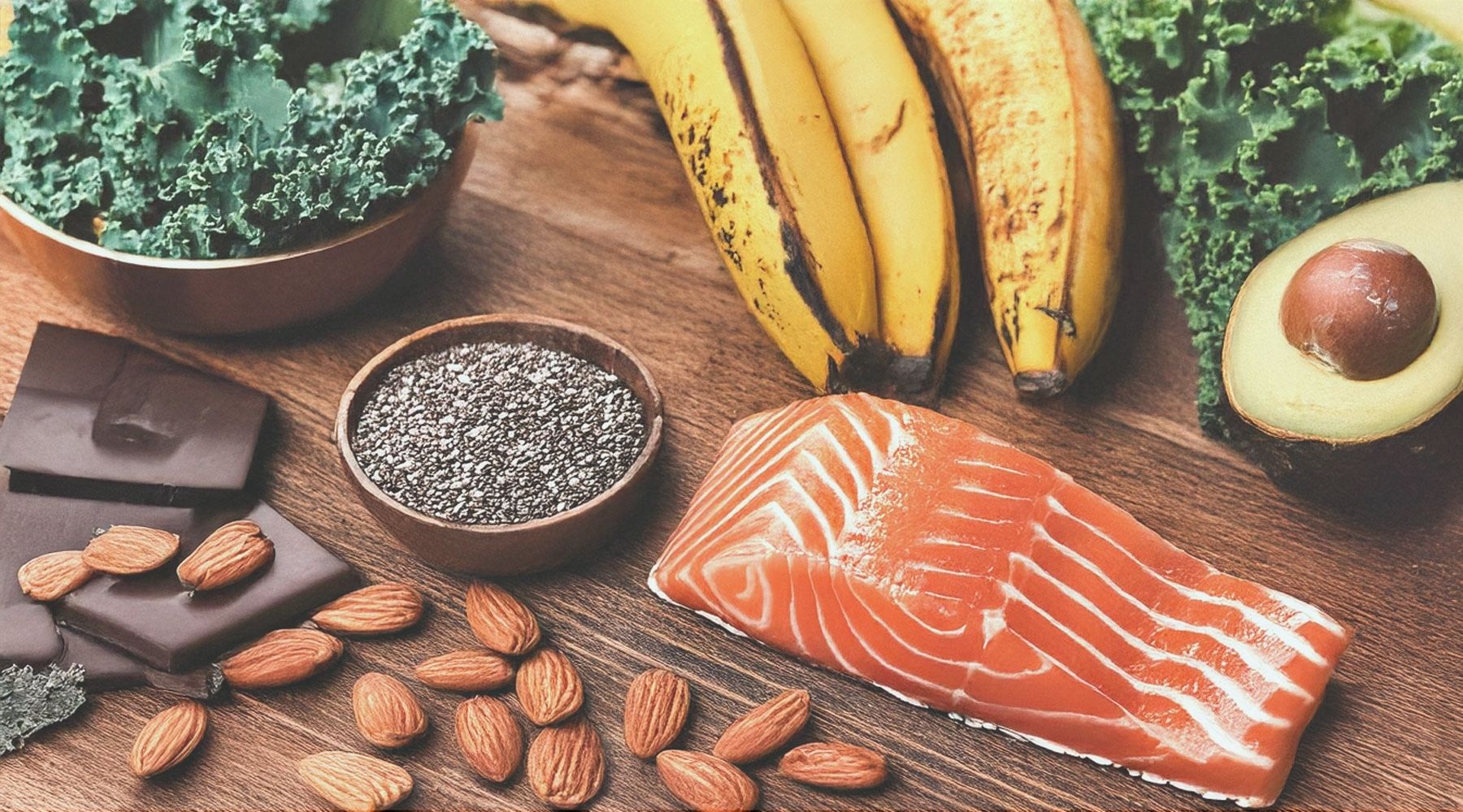 Electrolyte-rich foods including salmon, kale, chia seeds, almonds, dark chocolate, bananas, and avocado on a wooden table. These foods provide essential nutrients like potassium, magnesium, and omega-3 fatty acids.