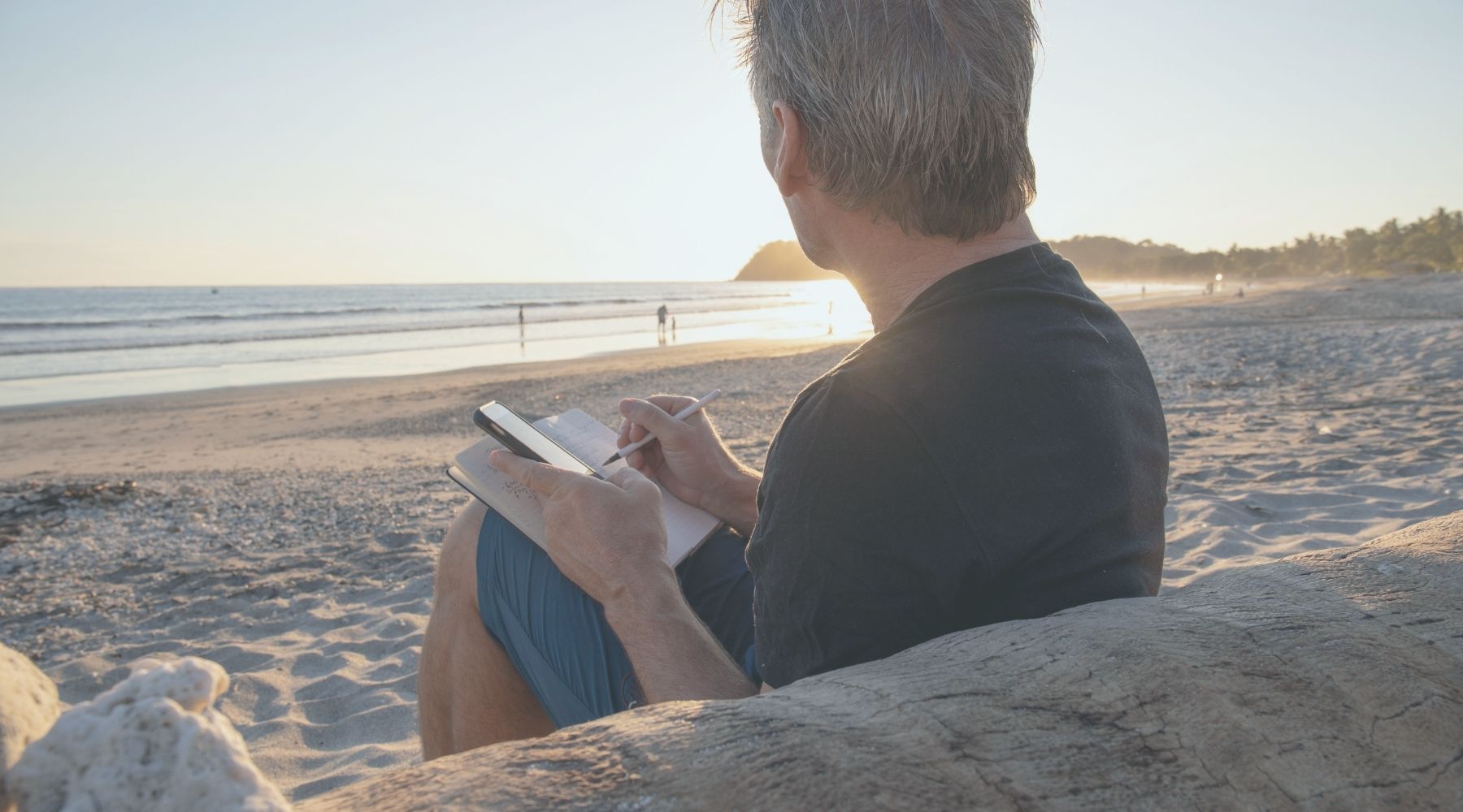 A man sitting on a beach journaling in a notebook during sunset, reflecting on his thoughts with the peaceful ocean in the background.