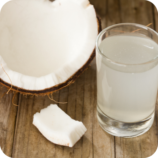 Fresh coconut water in a glass with coconut halves and meat on a rustic wooden surface.