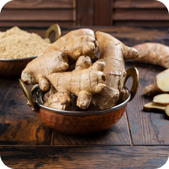 Fresh ginger roots in a copper bowl with ground ginger and sliced pieces on a wooden surface.