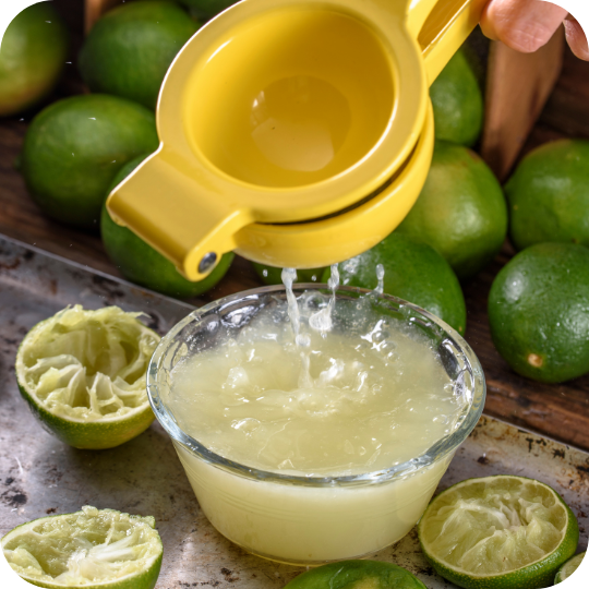 Fresh lime juice being squeezed into a bowl with whole and halved limes in the background.